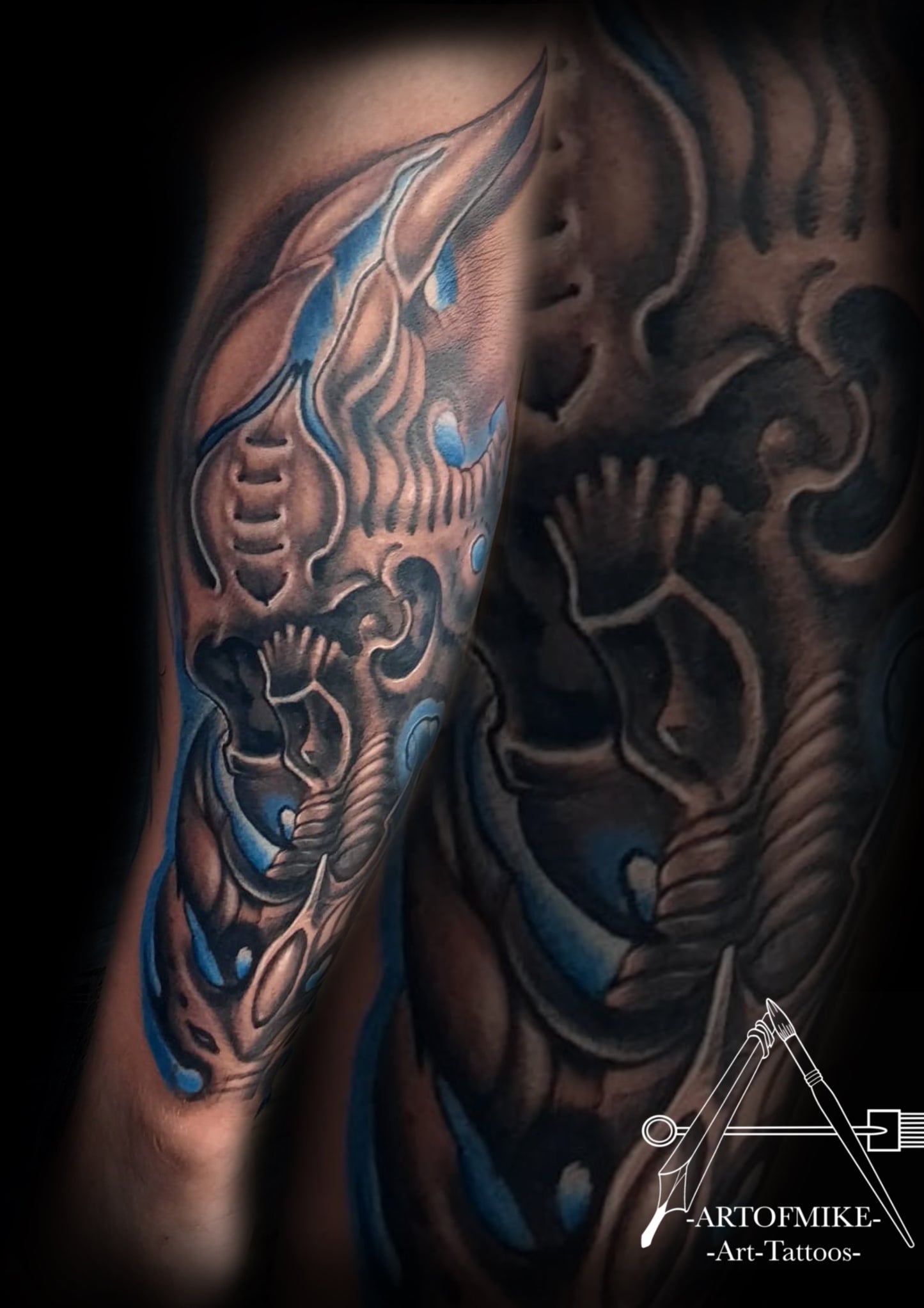 A forearm biomechanical tattoo with blue, gray, and black abstract shapes blends into the skin. Book your Tattoo now with Artofmike @innerdarkness at Artofmiketattoos.com; artist’s logo appears in the corner.